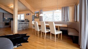 A modern interior with a wooden ceiling, a dining area with chairs and a bench, a kitchen with cabinets, and a large window with curtains.