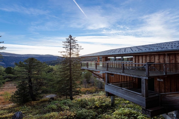A wooden cabin with a balcony overlooking a forested landscape and distant mountains.