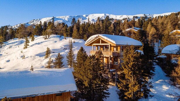 A wooden chalet with a balcony, surrounded by snow-covered trees and mountains in the background.