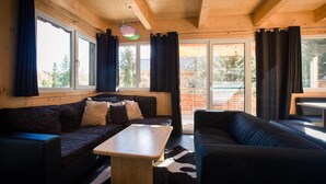 L’intérieur d’un chalet accueillant, avec des murs en bois, un canapé brun foncé, une petite table en bois et de grandes fenêtres ornées de rideaux.