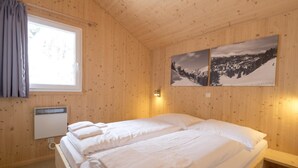 A wooden-paneled room with two beds, a window with curtains, a wall-mounted heater, and two framed pictures. - Charming chalet with indoor hot tub (Turracherhöhe)