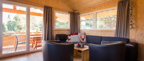 Interior acogedor de una cabaña con techo de madera, ventanas amplias, un sofá, dos sillones y una mesita.