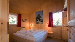 Un dormitorio de cabaña de madera con una cama, una mesita de noche, una ventana con cortinas rojas y un cuadro de un árbol.