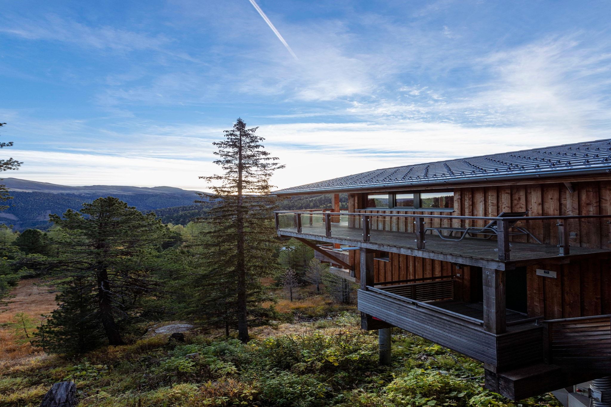 A wooden cabin with a balcony overlooking a forested landscape and distant mountains.
