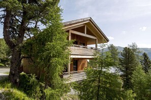 A two-story wooden cabin with a balcony, surrounded by trees and overlooking a lake.