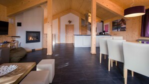 Ein geräumiger Wohnbereich mit Kamin, Holzbalken, einem Essbereich mit Stühlen und einer Küche im Hintergrund.