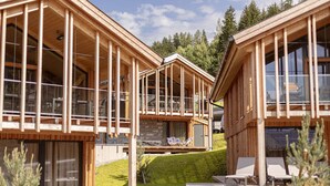 A wooden cabin with large windows and a balcony, surrounded by a grassy area and trees.