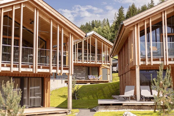 Una baita di legno con ampie finestre e un balcone, immersa nel verde, circondata da un prato e alberi.