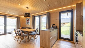 A modern kitchen with wooden cabinetry and a central island, large windows with curtains, and a dining area with a wooden table and chairs.