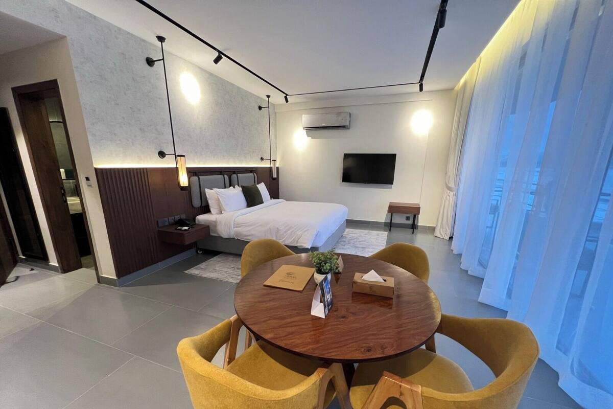 A modern hotel room with a bed, a TV, a small table with chairs, and a large window with curtains.