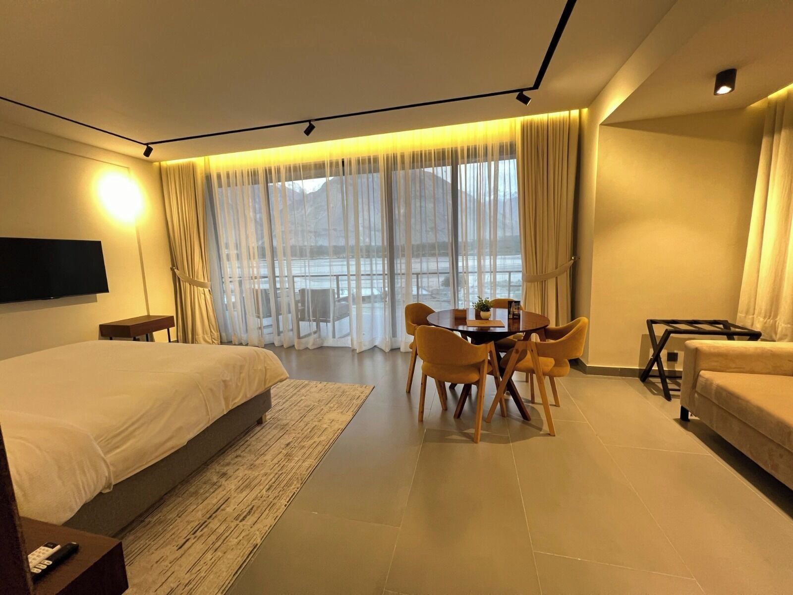 A hotel room with a large bed, a dining table with chairs, a sofa, and a TV mounted on the wall.
