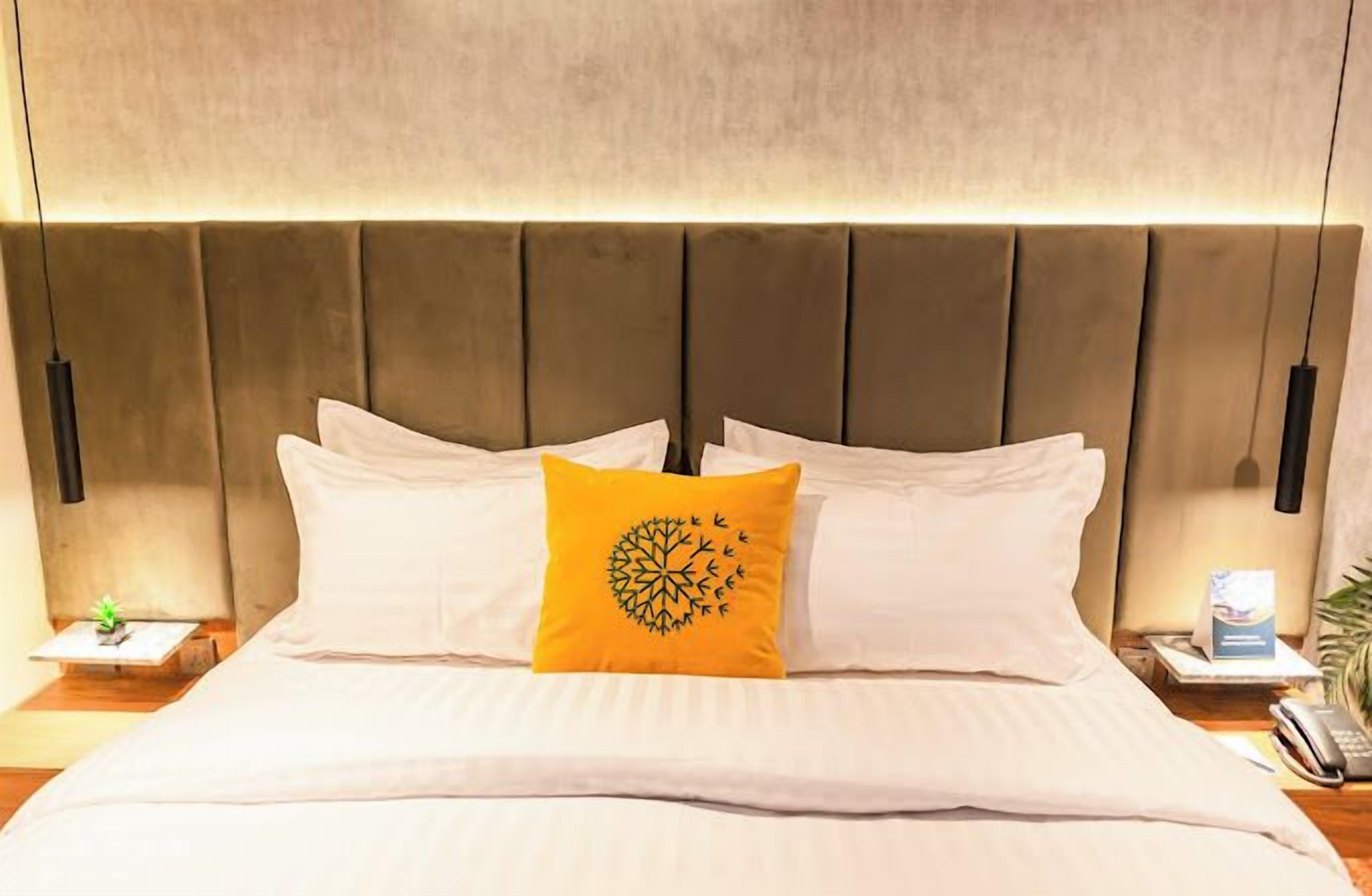 A hotel room with a large bed, a headboard with vertical panels, a nightstand with a lamp, and a small table with a plant.