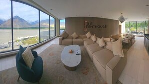 A spacious living room with a large window overlooking a mountainous landscape. The room features a beige sectional sofa, a round coffee table, and a teal armchair. The wall has a sign that reads "THE PENTHOUSE." - Himmel Skardu by Luxus (Wazirpur)