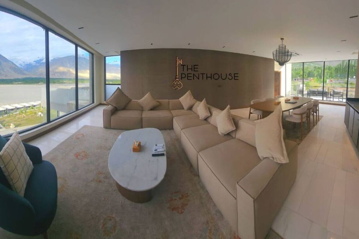 A spacious living room with a large window overlooking a mountainous landscape. The room features a beige sectional sofa, a round coffee table, and a teal armchair. The wall has a sign that reads "THE PENTHOUSE."