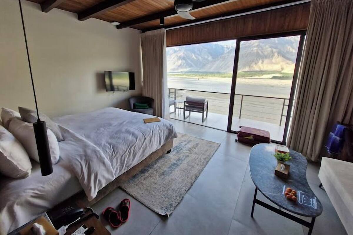 A modern hotel room with a large bed, a flat-screen TV, a balcony with a view of mountains, and a small table with a plant.