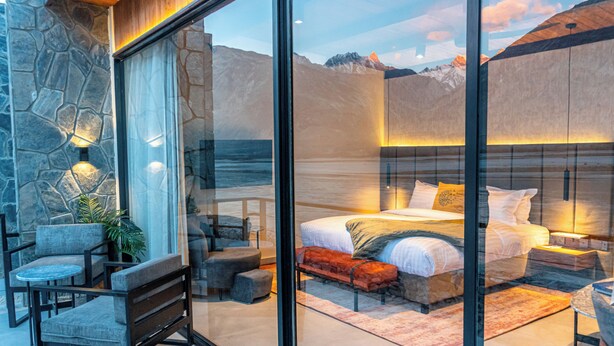 A modern bedroom with a stone wall, a large bed, a seating area with a bench and chairs, and a view of mountains through a glass door. - Himmel Skardu by Luxus (Wazirpur)