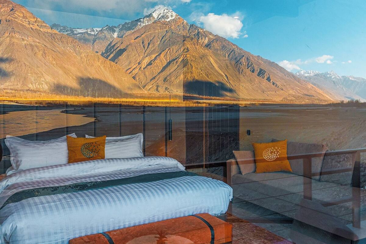 A bedroom with a bed, a glass table, and a chair with a view of mountains and a lake.