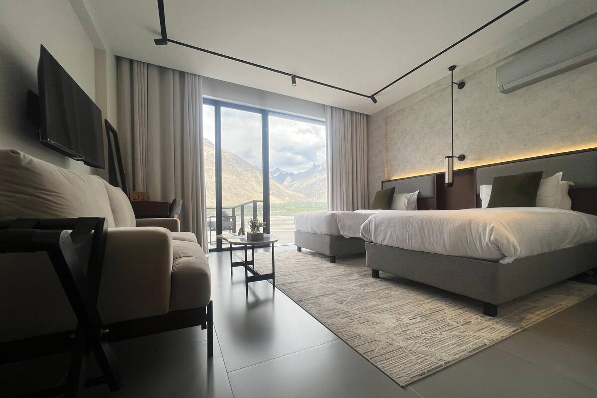 A modern hotel room with a large window, a sofa, two beds, and a flat-screen TV.