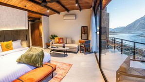 A modern hotel room with a large bed, a sofa, a coffee table, and a view of a mountainous landscape through a glass door. - Himmel Skardu by Luxus (Wazirpur)