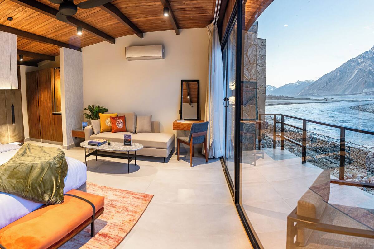 A modern hotel room with a large bed, a sofa, a coffee table, and a view of a mountainous landscape through a glass door.