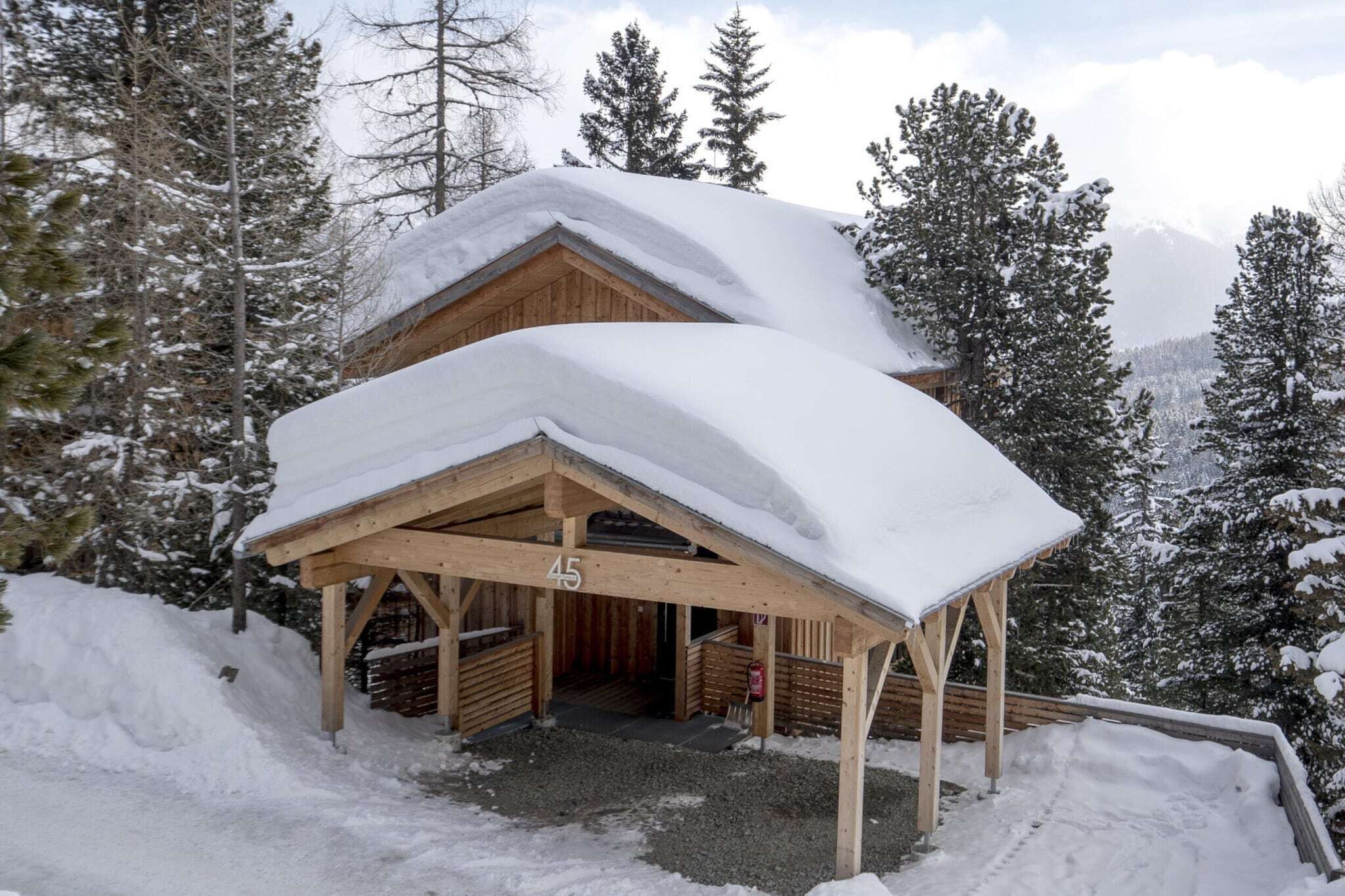 Um chalé de madeira com telhado coberto de neve, rodeado por árvores e montanhas também cobertas de neve.