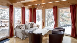 L’intérieur chaleureux d’un chalet, avec un canapé, des fauteuils et une terrasse en bois visible à travers de grandes fenêtres.