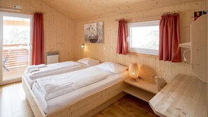 Une chambre dans un chalet en bois, avec deux lits, une fenêtre avec des rideaux, une lampe fixée au mur et une petite table.