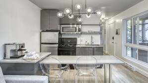 A modern kitchen with stainless steel appliances, a dining area with a clear glass table and chairs, and a large window providing natural light.