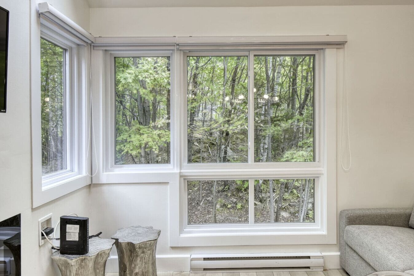 A room with a large window overlooking a forest, a small table with a decorative item, and a grey sofa.