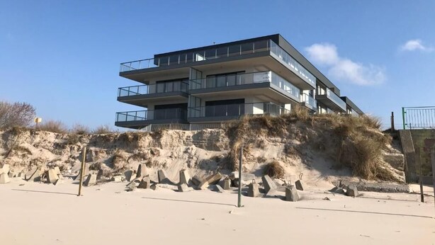 Un immeuble à plusieurs étages, doté de balcons, situé sur une plage de sable avec une falaise rocheuse en arrière-plan.