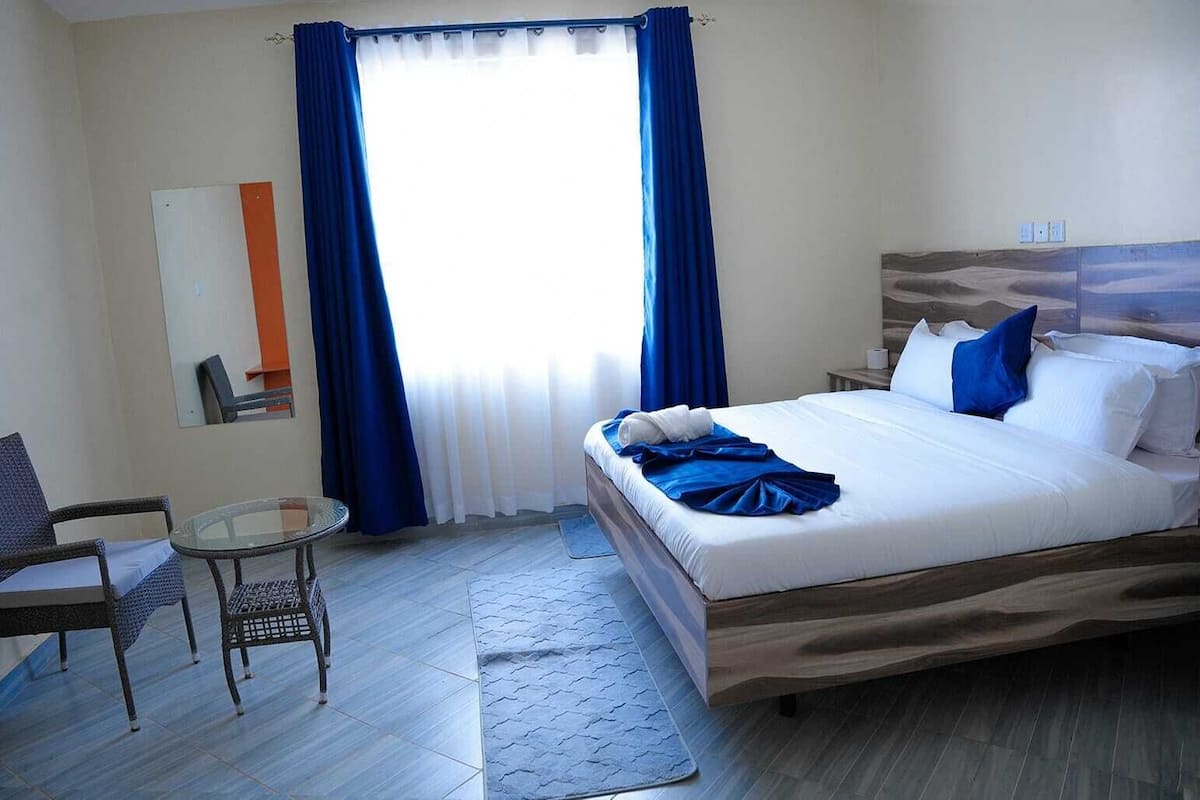 Quarto de hotel com uma cama, uma cadeira, uma mesinha e uma janela com cortinas azuis.
