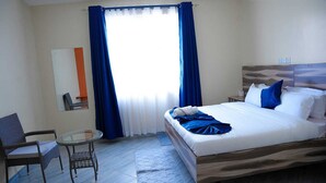 A hotel room with a bed, a chair, a small table, and a window with blue curtains.