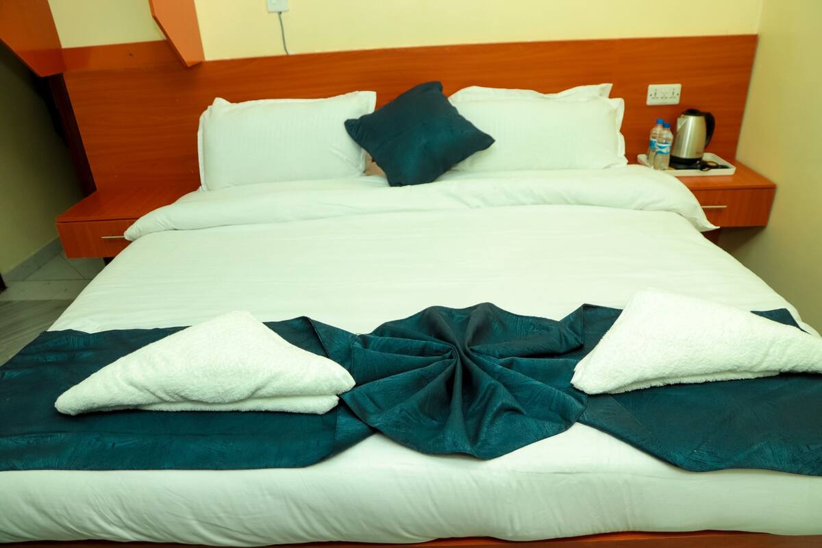 Uma cama bem arrumada com roupa de cama branca e azul-turquesa, uma cabeceira de madeira e uma mesa de cabeceira com chaleira e garrafa de água.
