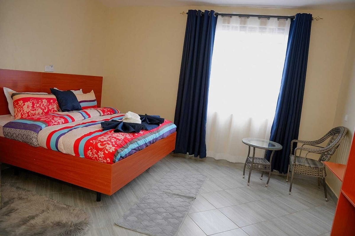 Quarto de hotel com cama de madeira, uma mesa redonda pequena e uma cadeira.