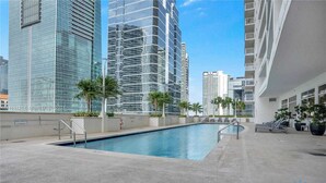 A rooftop pool with a view of high-rise buildings, surrounded by palm trees and lounge chairs. - Beautiful 1BR Apartment with Amenities (Miami)