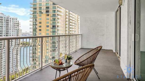 A balcony with wicker chairs, a small table with a bottle and fruits, and a view of high-rise buildings and a river. - Beautiful 1BR Apartment with Amenities (Miami)