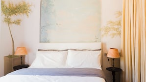 Een slaapkamer met een groot, rondboogvormig hoofdbord, twee nachtkastjes met lampen, een bed met wit en grijs beddengoed, en een muurschildering van een boom.