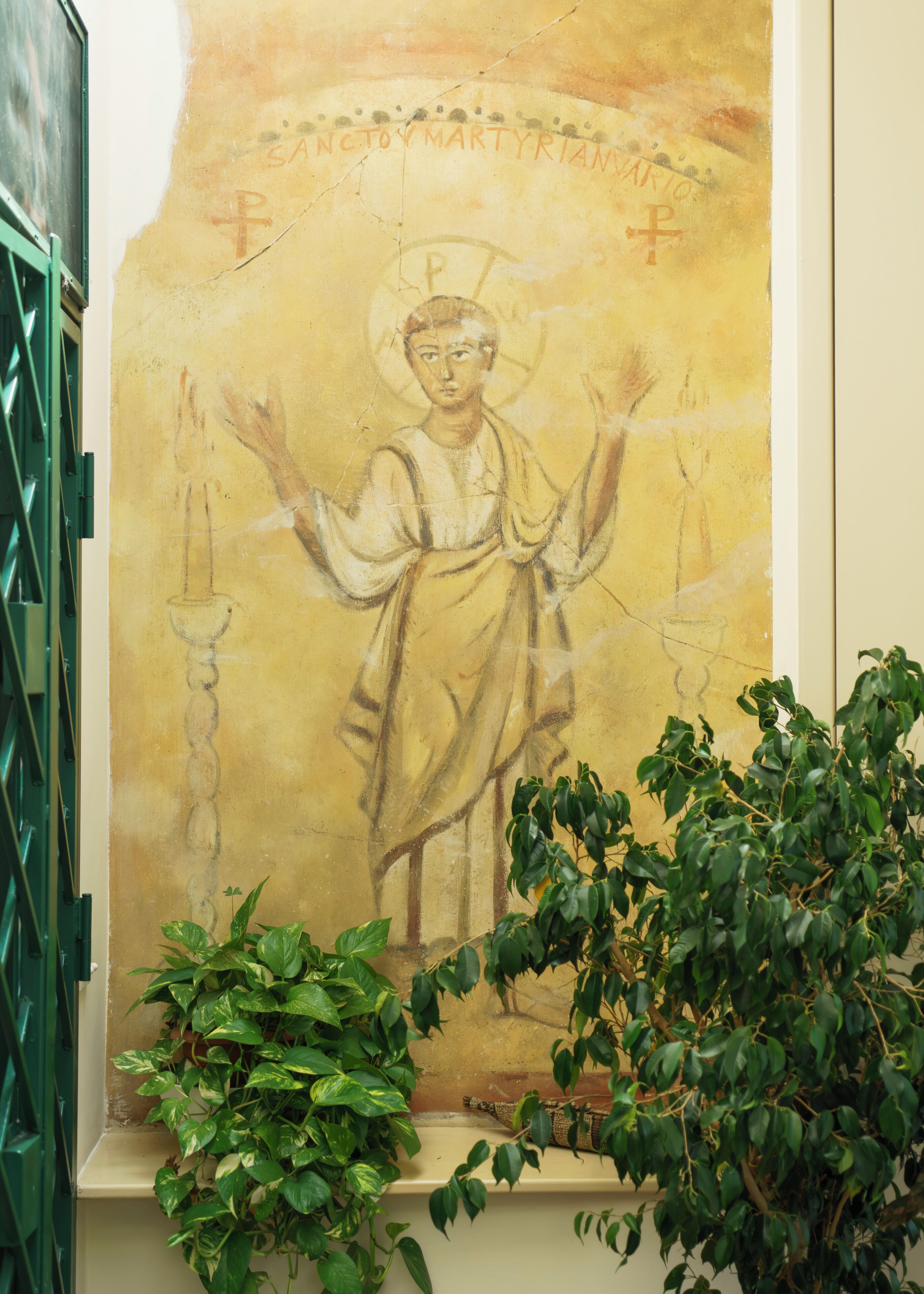 A wall with a religious icon, a green metal gate, and green foliage.