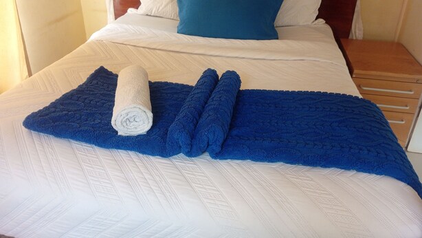 A neatly made bed with a blue blanket and rolled towels, a wooden headboard, and a nightstand with drawers. - Lotus House Harare (Harare)