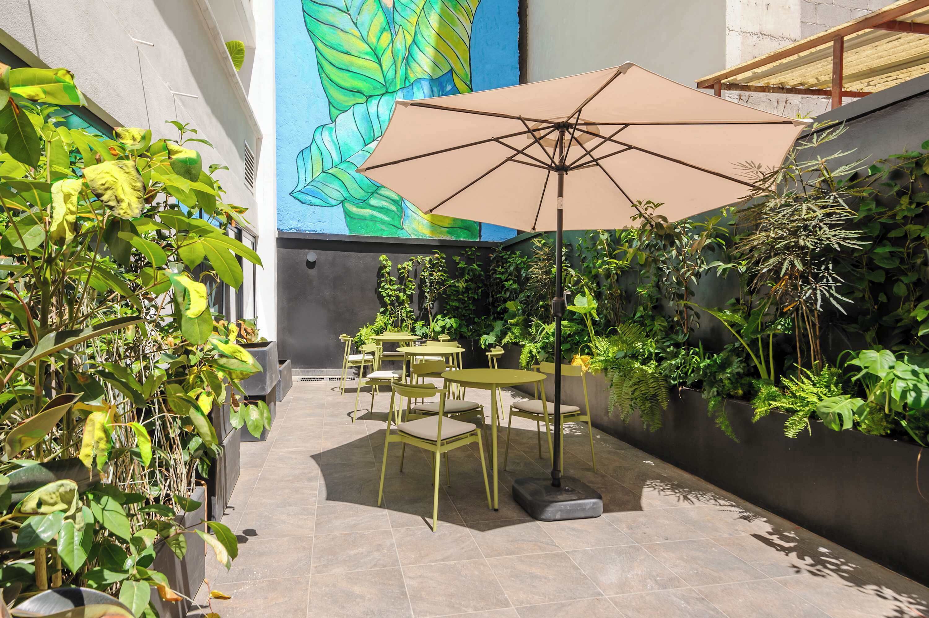 An outdoor patio with green chairs and a large umbrella, surrounded by lush plants and a colorful mural.