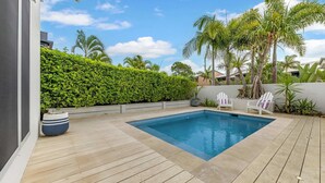 A swimming pool with a wooden deck, surrounded by a hedge and palm trees.