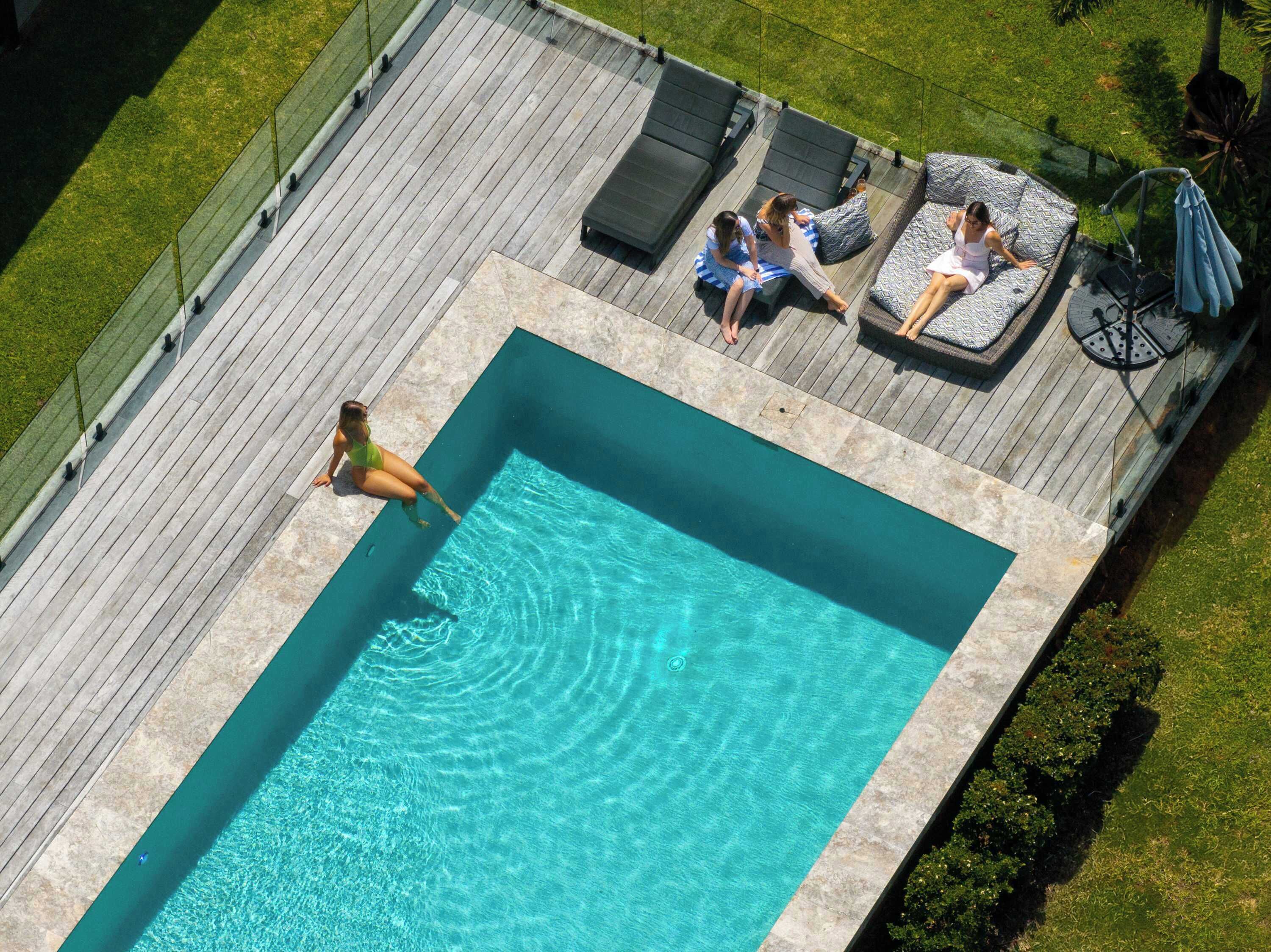 A rectangular swimming pool with a wooden deck surrounding it. There are lounge chairs and a small table with umbrellas on the deck.