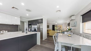 A modern kitchen with a central island, a dining area with white chairs, and a living space with a sofa and a coffee table. - Family Home in Maroochydore (Maroochydore Region)