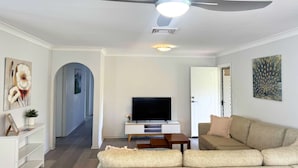 A living room with a ceiling fan, a flat-screen TV on a white entertainment unit, a beige sofa, and a hallway leading to another room. - Family Home in Maroochydore (Maroochydore Region)
