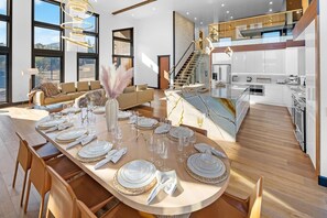 Una cocina moderna con una isla central, un comedor con una mesa redonda dispuesta para seis personas, y una escalera que conduce a un nivel superior.