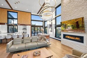 A modern living room with a sofa, a dining area with a table, and a fireplace. - Sky View Manor: Luxurious! Oversized Swim Spa/Hot Tub! Newly Built! Chef’s Kitchen! Smart Home/High Tech! Outdoor Fire Pit! Massive Deck! Forest Walks! Game & Entertainment Room w/ Pool Table, Poker Table & Bar! (Big Bear City)