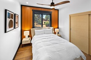 Un dormitorio con cama, dos mesitas de noche, ventilador de techo, ventana con vistas y cuadros en la pared.