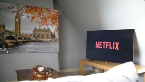 A room with a television displaying the Netflix logo, a wall-mounted picture of a bridge and buildings, and a wicker basket on a table. - Georgianstyle-2bed-smarttv-freeparking (Boston)