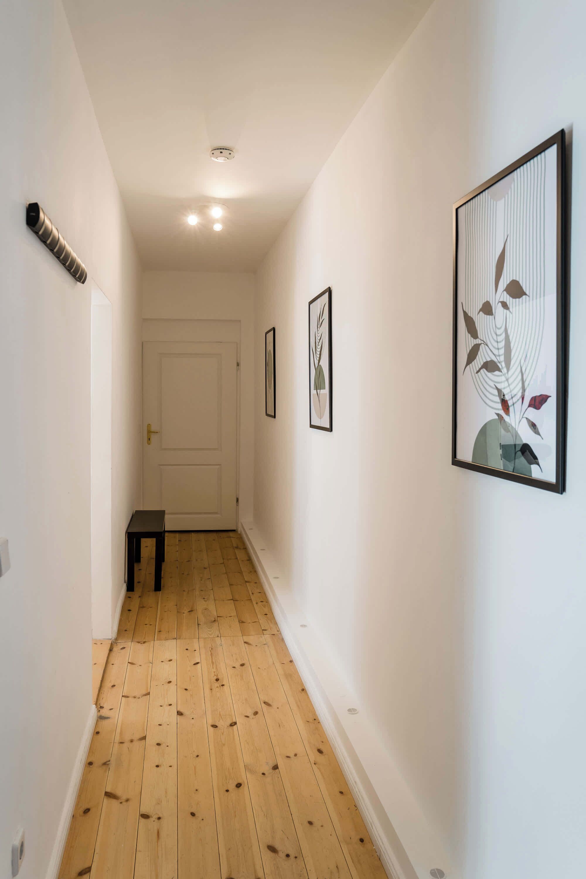 Un pasillo estrecho con suelo de madera, paredes blancas y cuadros enmarcados.
