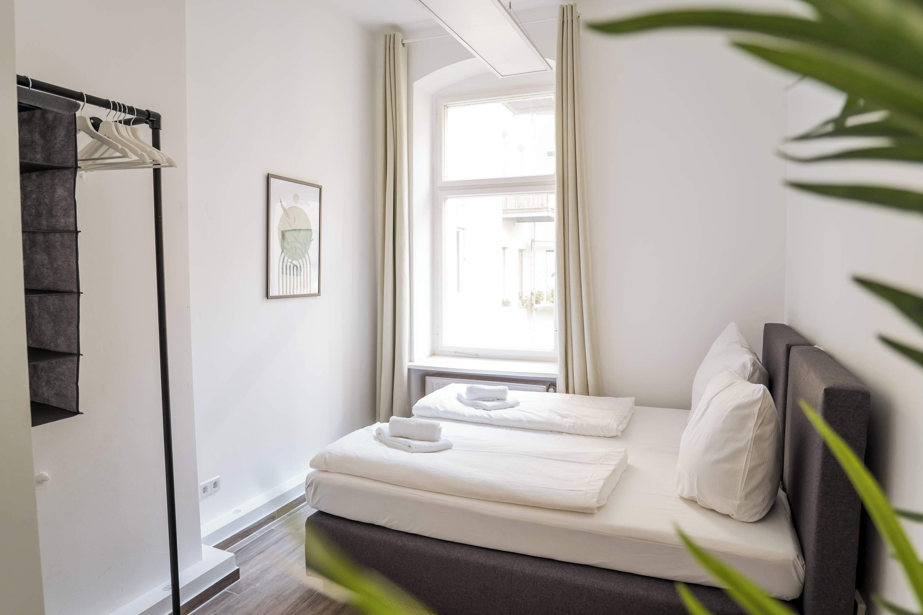 A neatly made bed with white linens and pillows, a window with curtains, and a wall-mounted towel rack.